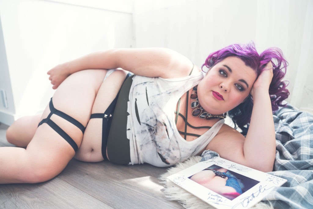 Woman with purple hair lounging on hardwood floor among flannel shirts.