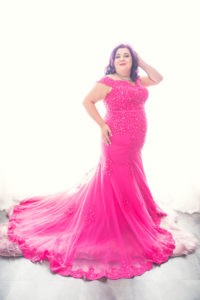 Woman with purple hair in long bright pink gown against a light wall.