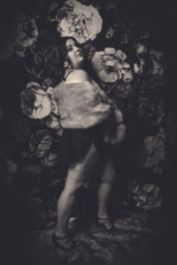 Black and white photo of a woman with long dark hair wearing a short black dress and fur stole against a floral backdrop.