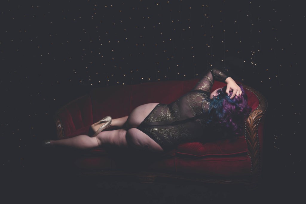 Woman lounging on red couch facing wall wearing glittery bodysuit and gold heels.