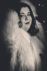 A black and white headshot of a woman with dark hair and lipstick wearing a furry coat.