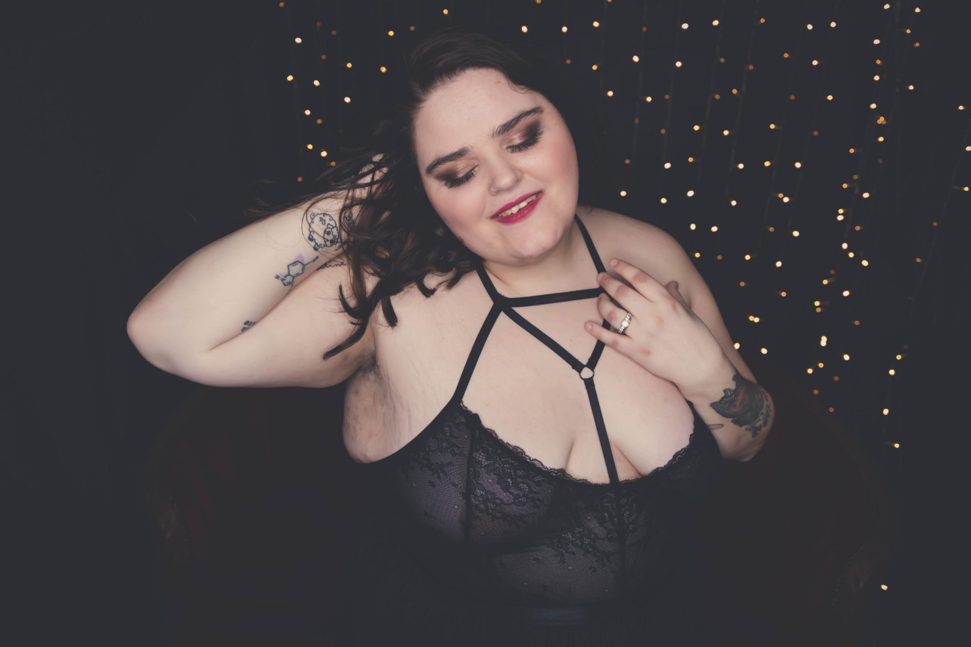 Woman with long dark hair, tattoos and scrappy lingerie stands with her eyes closed in front of twinkly lights.