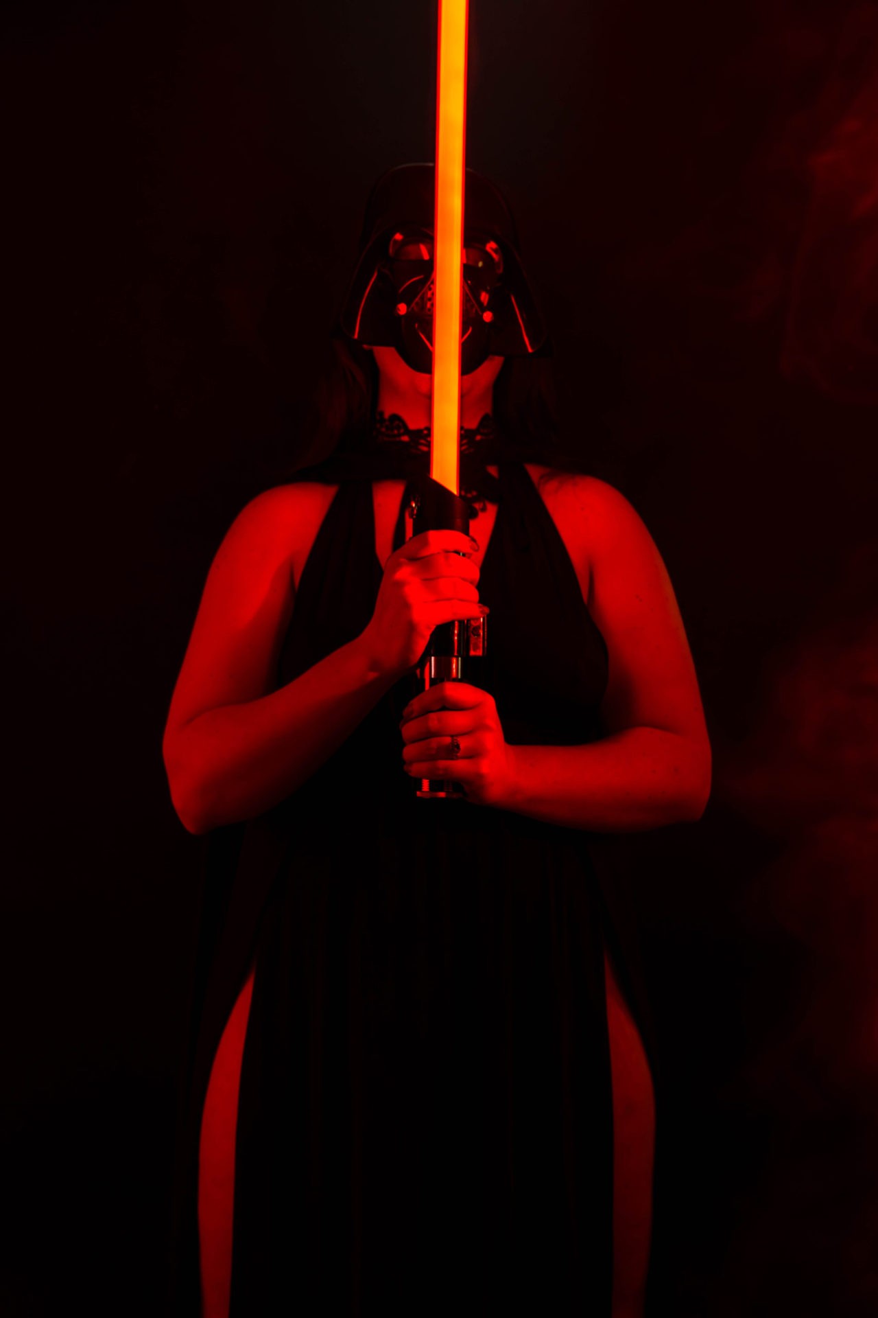color image of a woman holding a red light saber under a red light with a black background wearing a Darth Vader helmet.